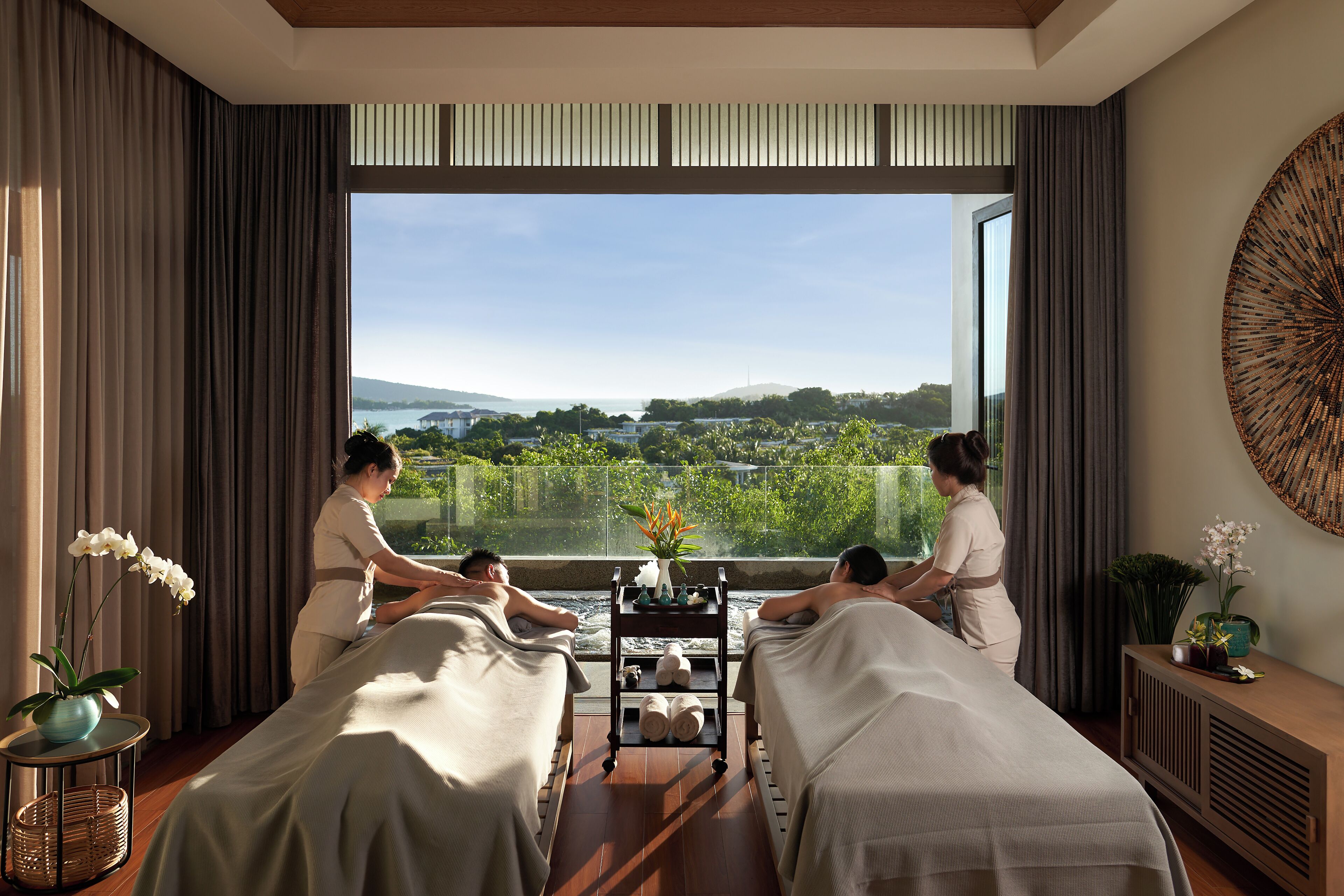 Un cuarto de spa con dos camillas de masaje, un ventanal con vistas al paisaje, y una mesita con un florero.