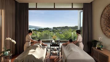 Un cuarto de spa con dos camillas de masaje, un ventanal con vistas al paisaje, y una mesita con un florero.