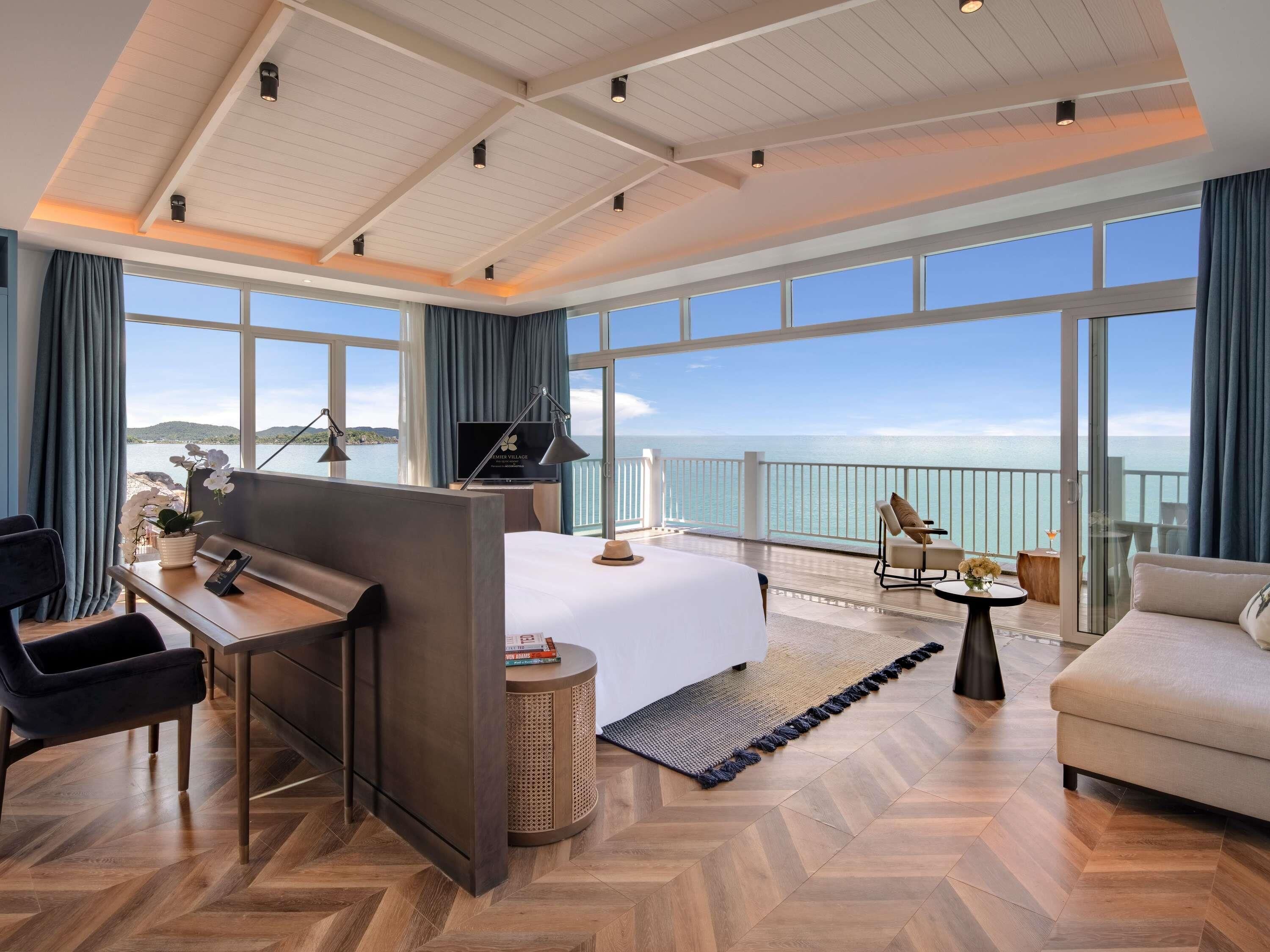 A modern hotel room with a large bed, a desk, a chair, and a sofa. There is a large window with a view of the sea.