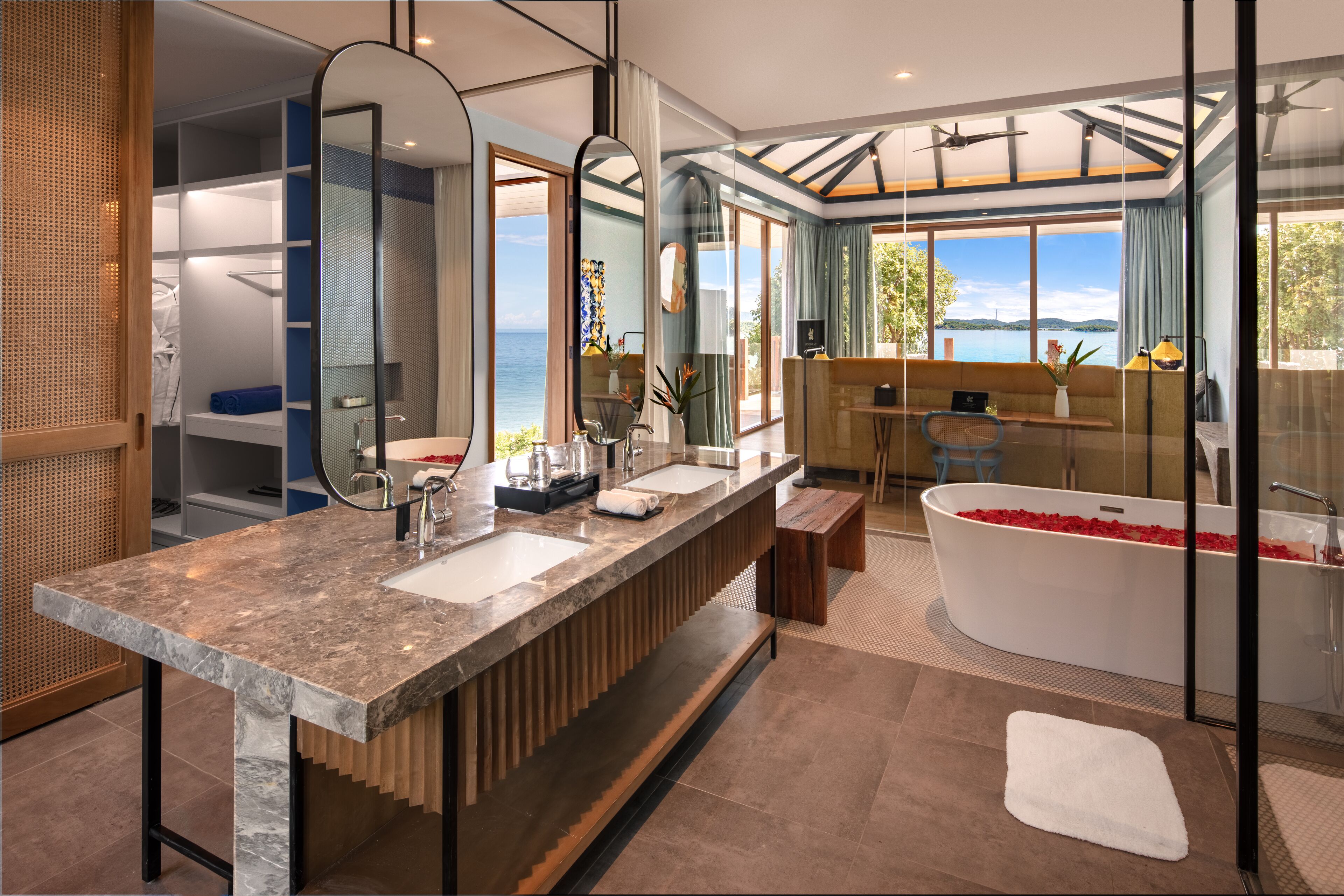 A modern bathroom with a large vanity, dual sinks, a freestanding bathtub, and a view of the sea.