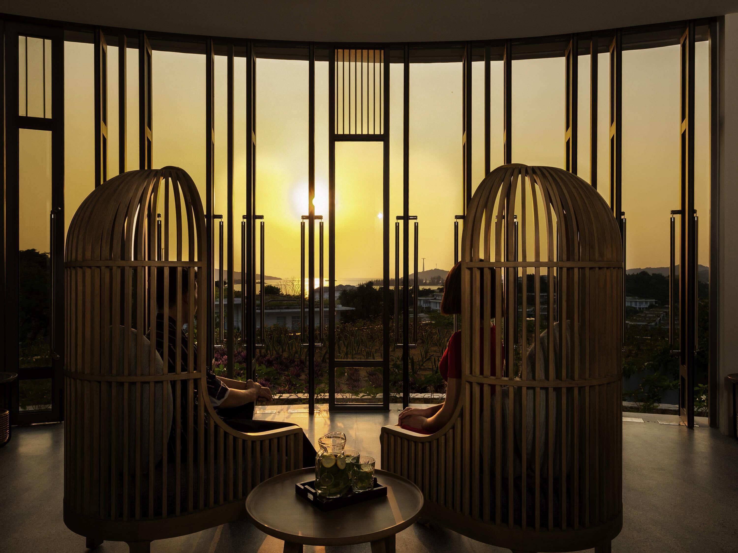 A room with two wooden-framed chairs facing a large window with vertical blinds, a small round table with a tray, and a view of a distant landscape.