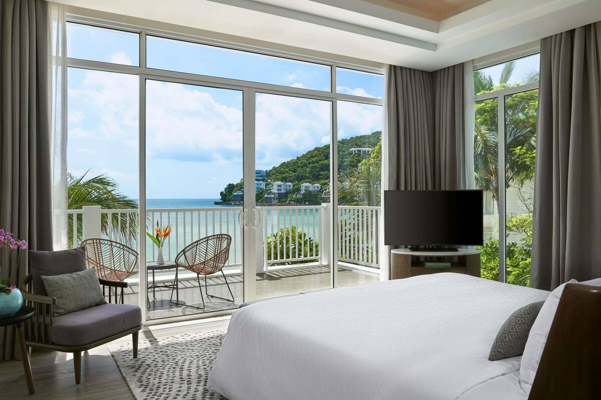 Une chambre d’hôtel dotée d’un grand lit, d’une télévision, d’un balcon avec vue sur la mer et d’un coin salon avec des fauteuils.