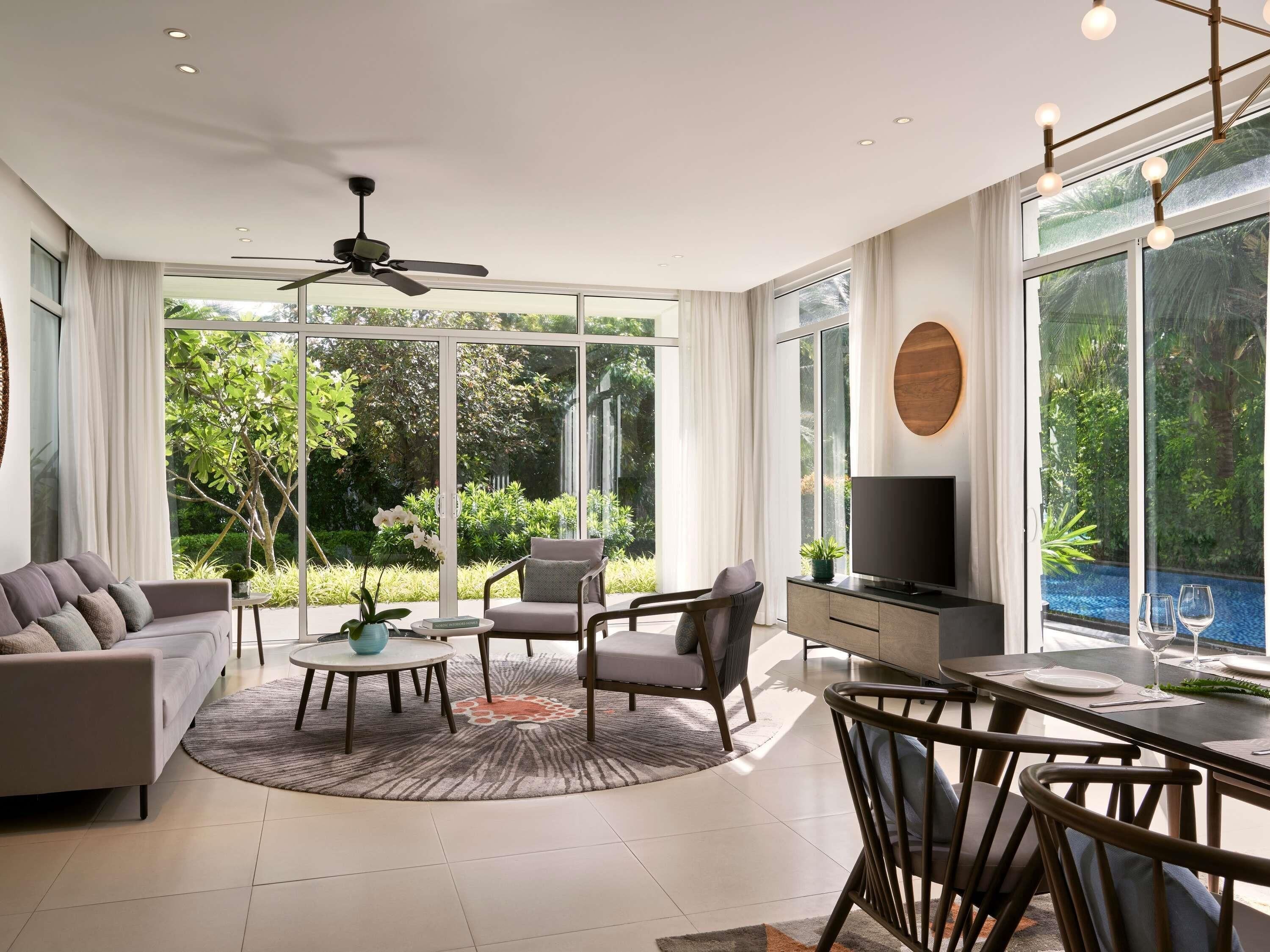 A modern living room with a sofa, chairs, a dining table, a TV, and large windows offering a view of greenery.