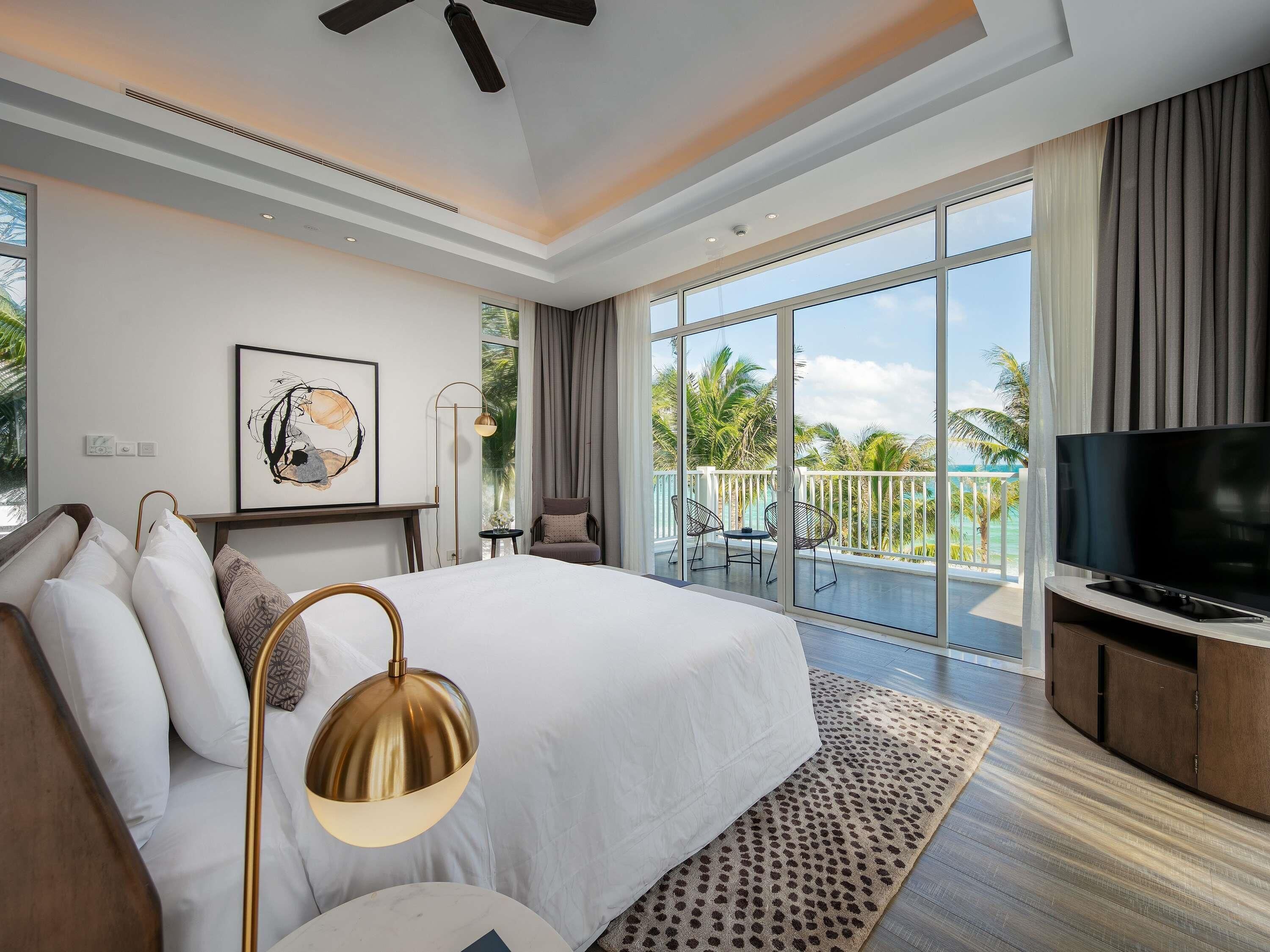 A hotel room with a bed, a TV, a balcony with a view of palm trees, and a ceiling fan.