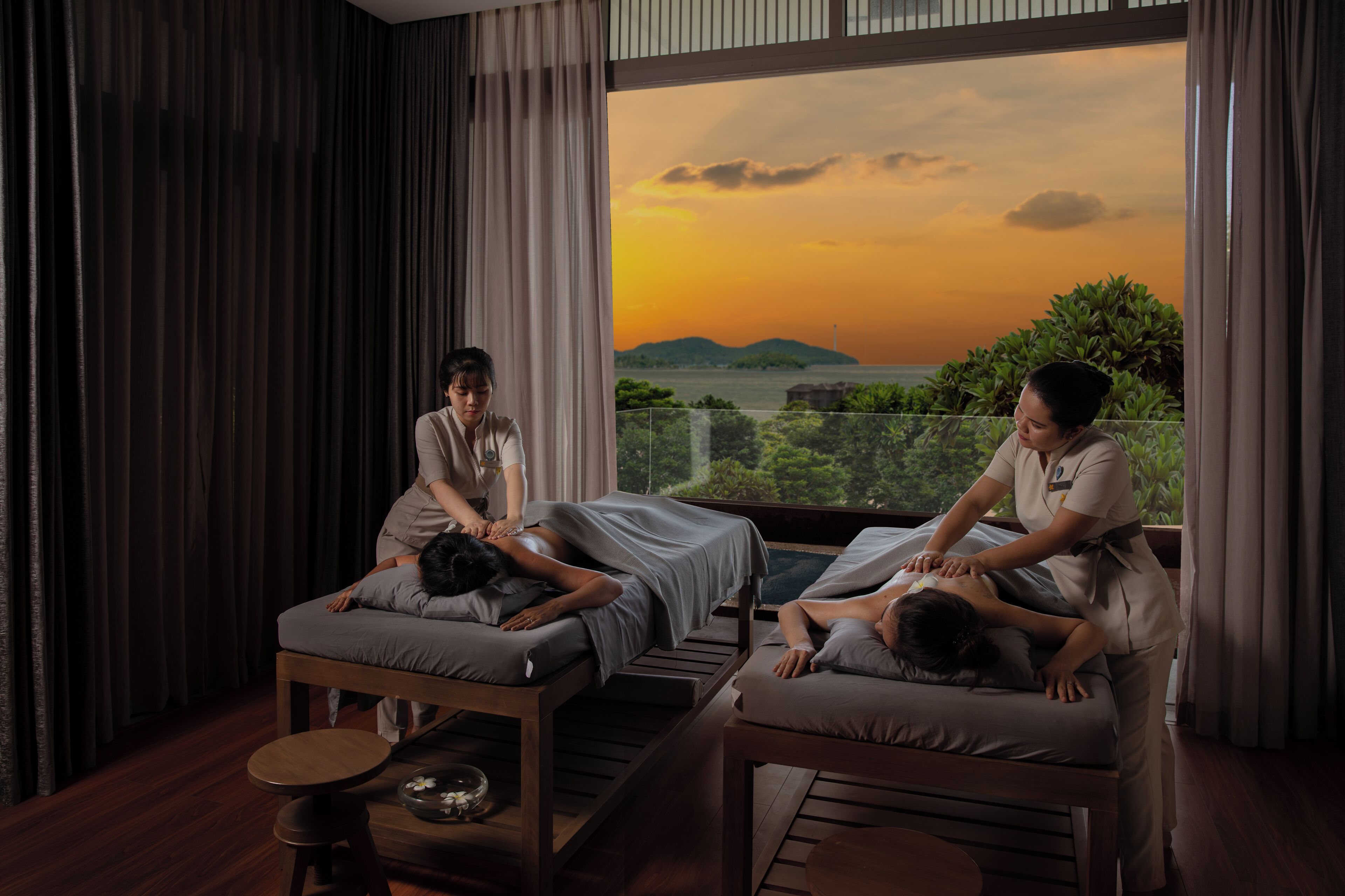 A spa room with two massage tables, a large window with a sunset view, and a wooden floor.