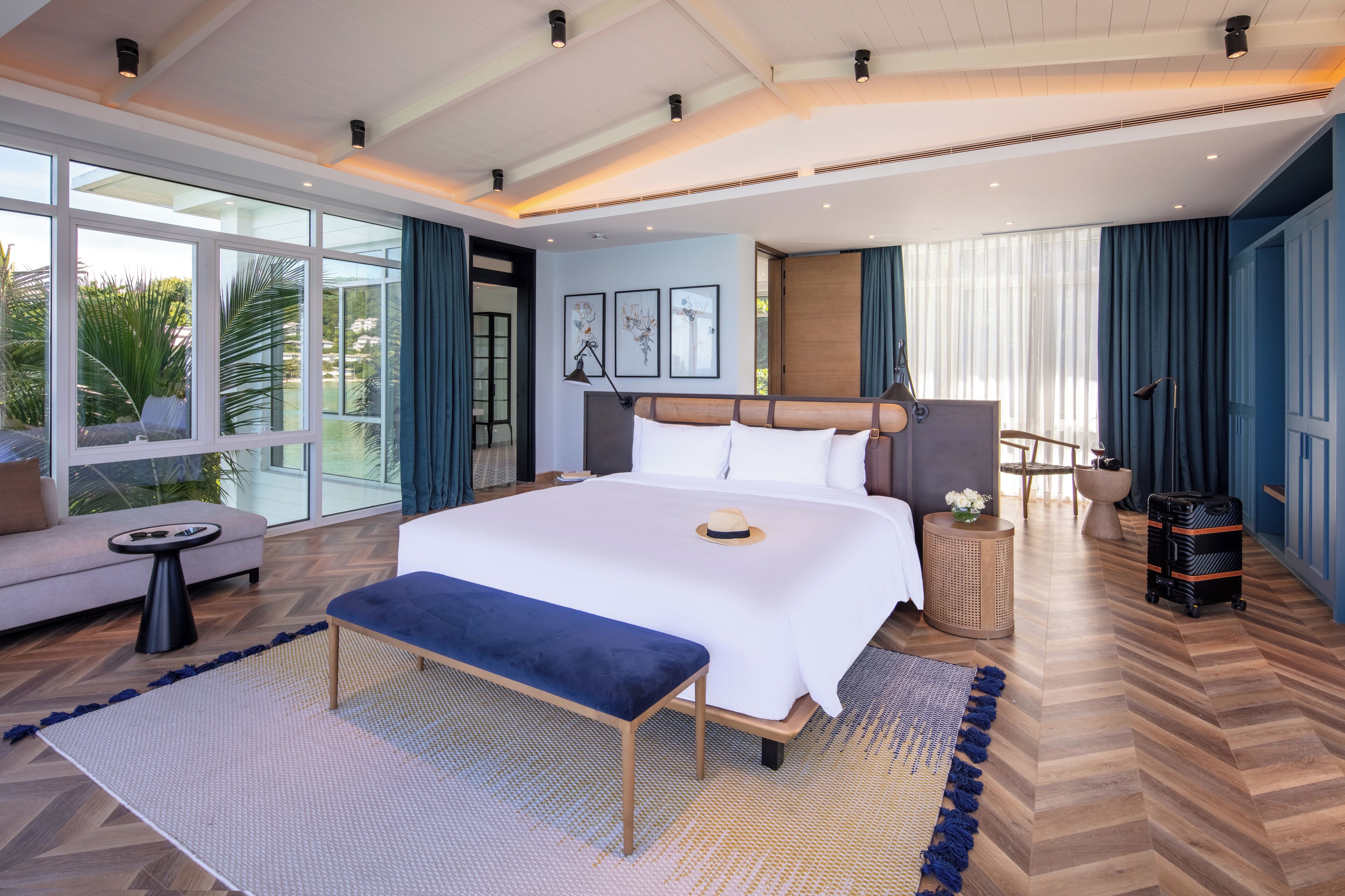 A modern hotel room with a large bed, a bench, a small table, and a view of the outdoors.