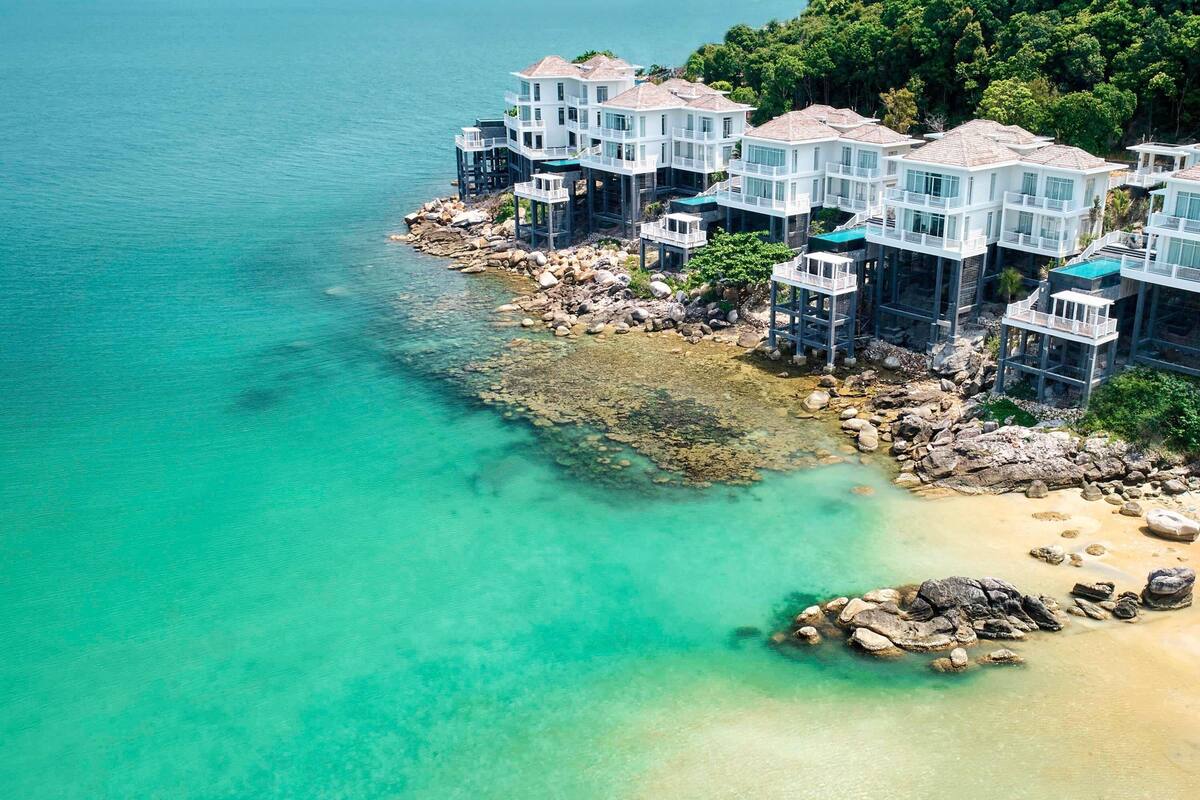 Une propriété située sur la côte, composée de plusieurs bâtiments blancs, dont certains sont dotés de balcons, et bordée d’une côte rocheuse aux eaux turquoise et cristallines.