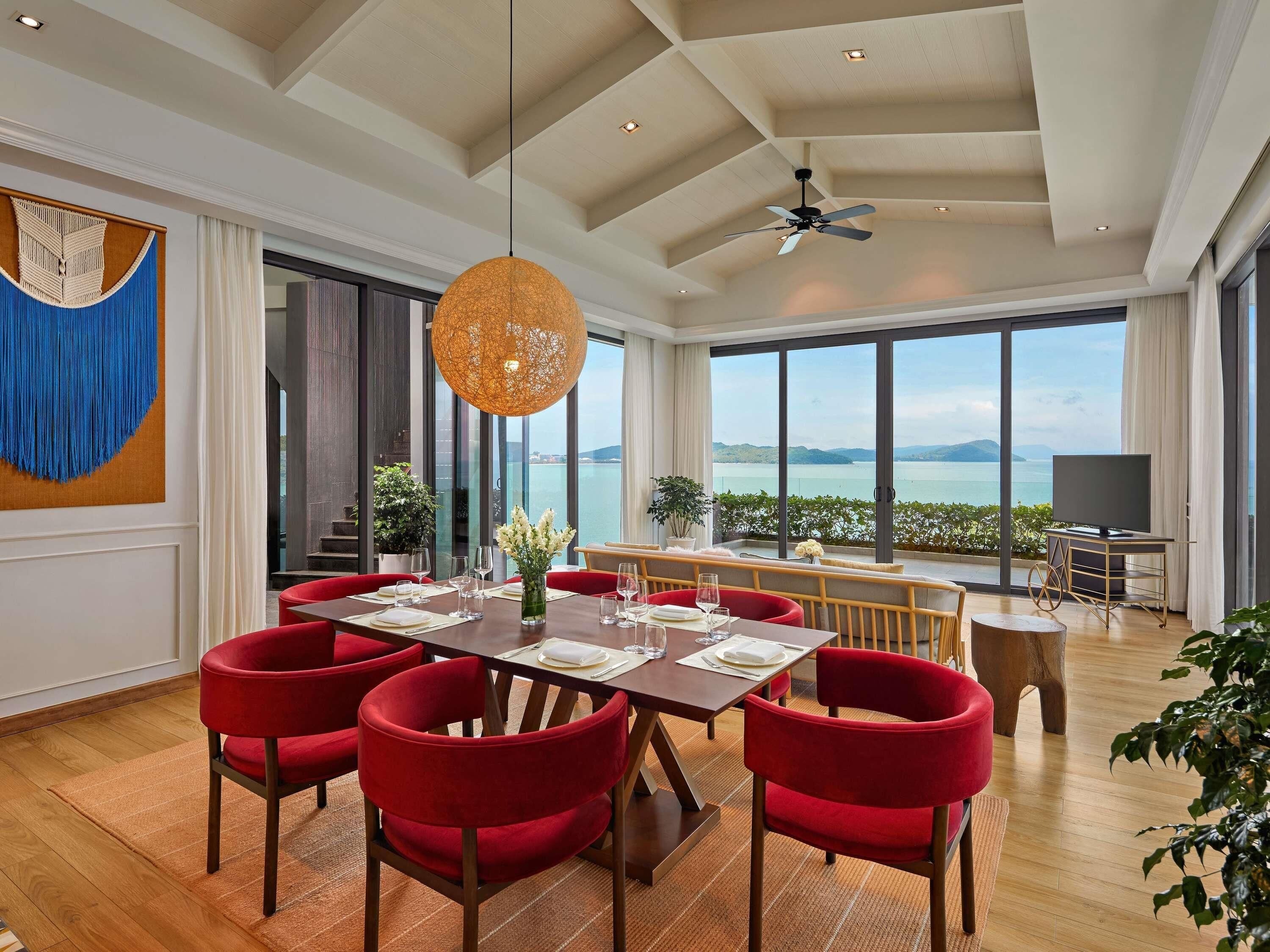 A dining area with a table set for six, red chairs, a ceiling fan, and a large window with a view of the sea.