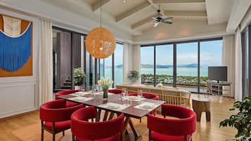 A dining area with a table set for six, red chairs, a ceiling fan, and a large window with a view of the sea.