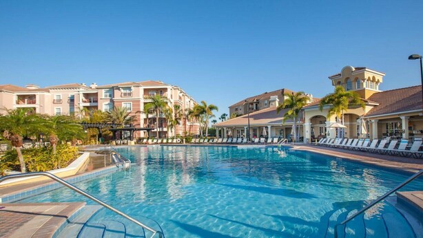 A swimming pool with lounge chairs and palm trees, surrounded by multi-story buildings with balconies.