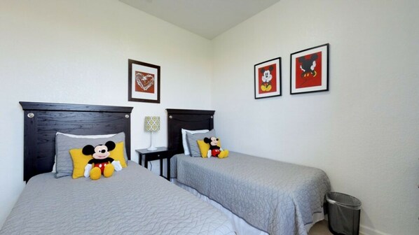 A hotel room with two beds, each with a Mickey Mouse plush toy and matching pillows. There are framed pictures on the wall, a small table with a lamp, and a trash bin. - Apartment in the Heart of Orlando BVD406 (Orlando)