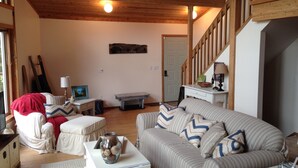 A cozy living room with a wooden ceiling, a sofa with striped cushions, a coffee table with a decorative jar, and a staircase with wooden railings. - Stunning Lake Views, Perfect U.P. Day-Trip Location, Day-Trip To Mackinac Island (St. Ignace)