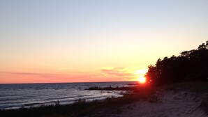 A beach at sunset with the sun setting on the horizon, sandy dunes, and sparse vegetation. - Stunning Lake Views, Perfect U.P. Day-Trip Location, Day-Trip To Mackinac Island (St. Ignace)