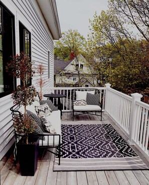 Un porche abrité avec une balustrade blanche, un banc en métal noir, une petite table et un tapis à motifs.