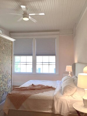 Une chambre à coucher avec un lit, un ventilateur de plafond, une fenêtre avec des stores, une lampe et un papier peint à motifs sur le mur.