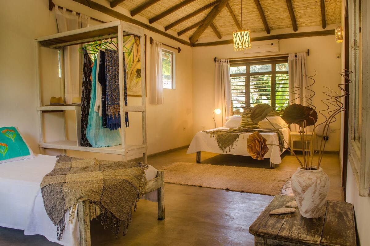 Une chambre rustique comprenant un lit, un banc en bois, un portant à vêtements et une grande fenêtre avec des rideaux.