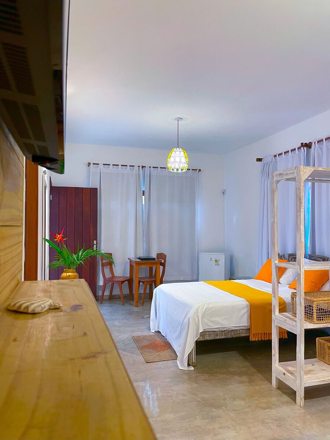 Ein Zimmer mit einem Bett, einer hölzernen Bank, einem Tisch mit Stühlen, einem kleinen Tisch mit einer Pflanze und einer Pendelleuchte.