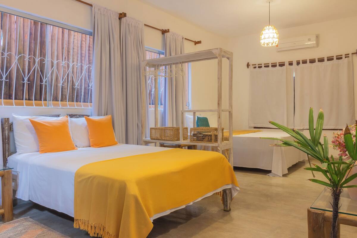 Une chambre d’hôtel avec un lit, une petite table, une chaise, un fauteuil suspendu et une plante en pot.