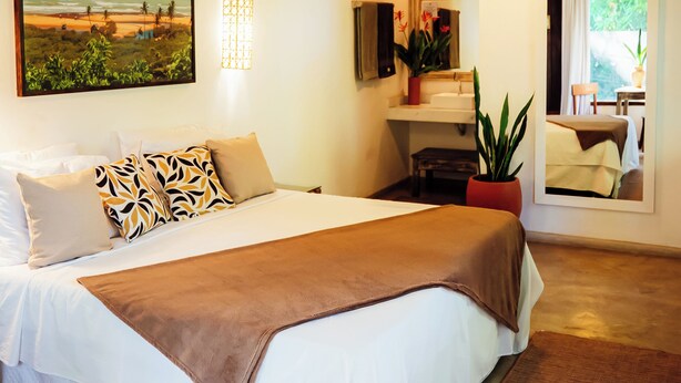 Une chambre à coucher avec un grand lit, un tableau représentant une plage, une plante en pot et un miroir qui reflète la pièce.