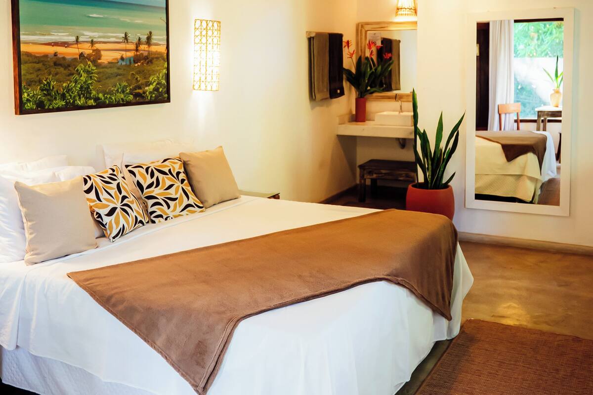 Une chambre à coucher avec un grand lit, un tableau représentant une plage, une plante en pot et un miroir qui reflète la pièce.