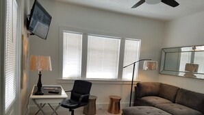 A room with a desk, office chair, and a large grey sofa. There is a window with blinds, a ceiling fan, and a floor lamp.