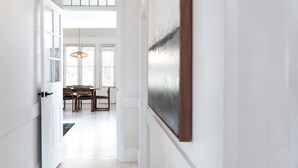 Un couloir mène à une salle à manger où se trouvent une table et des chaises, un luminaire suspendu et une œuvre d’art encadrée au mur.