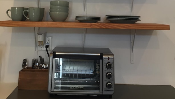 Une cuisine avec une horloge murale, des étagères remplies de vaisselle, un grille-pain et une fenêtre agrémentée d’un objet décoratif.