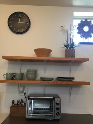 Una cocina con un reloj de pared, estantes con vajilla, un tostador y una ventana con un adorno.