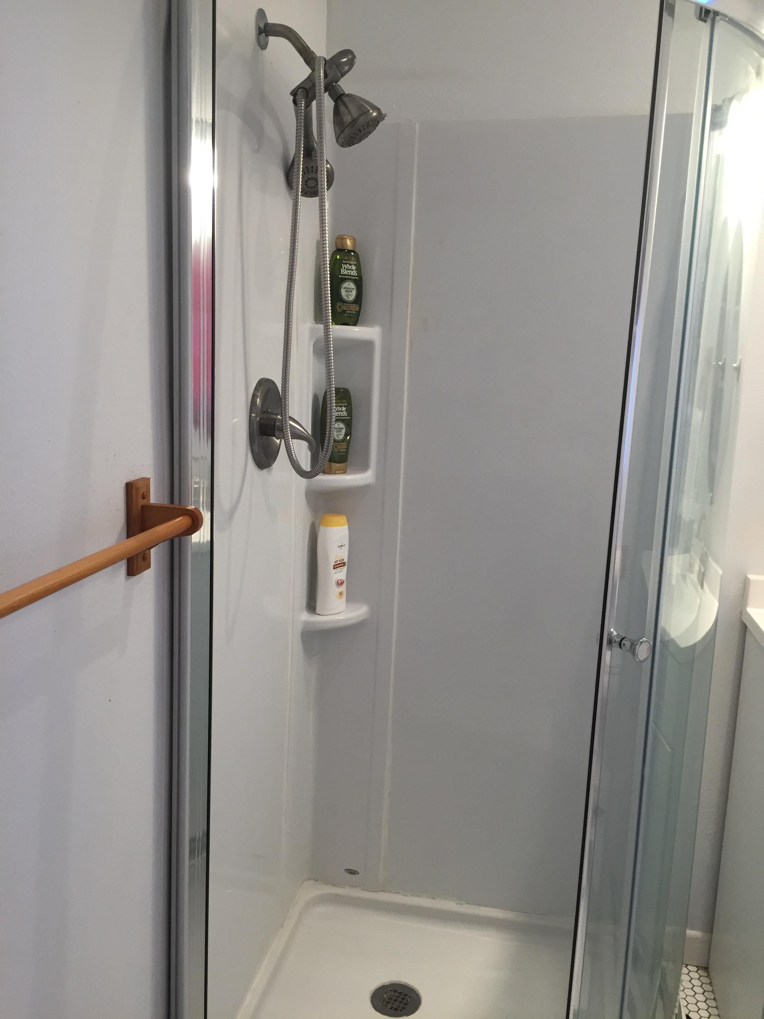 Eine Duschkabine mit Glaswänden, einer Handbrause und einem Handduschteil, sowie zwei Ablagen für Toilettenartikel.