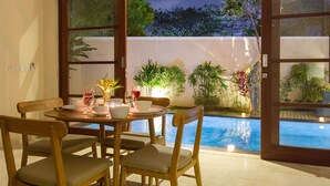 A dining area with a table set for four, surrounded by wooden chairs with cushions. A small table with a vase of flowers and wine glasses is in the foreground. The room has large glass doors leading to a pool area with a view of a garden and a statue. - 1BR Villa, DAILY HOUSE KEEPING! - W/Swimming Pool! (Seminyak)