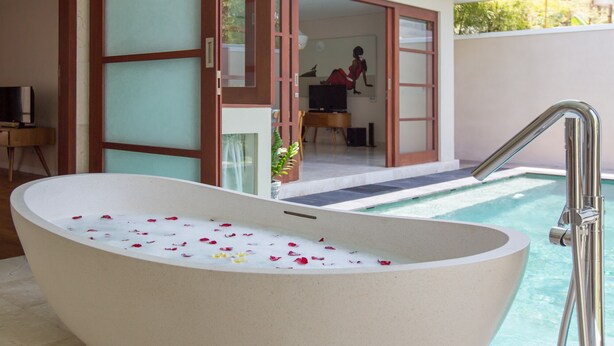 A modern bathroom with a freestanding bathtub filled with water and rose petals, a shower area, and a view of the living room through an open door. - 1BR Villa, DAILY HOUSE KEEPING! - W/Swimming Pool! (Seminyak)