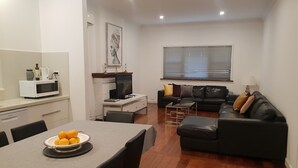 Un salon moderne avec un canapé en cuir noir, une table à manger sur laquelle se trouve une assiette d’oranges, un micro-ondes blanc et un téléviseur posé sur un meuble blanc.