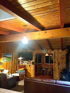 L’intérieur chaleureux d’un chalet avec un plafond en bois, un téléviseur, une bibliothèque et un coin repas.