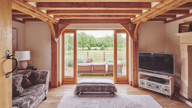 Ein gemütliches Wohnzimmer mit Holzbalkendecke, einer Couch, einem Fernseher auf einem Holzschrank und Blick auf einen Garten durch Glastüren.