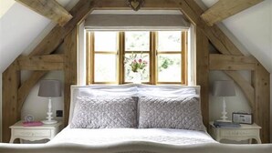 Ein Schlafzimmer mit einem Bett unter einer geneigten Holzbalkendecke, einem Fenster mit Blick auf Grünflächen und zwei Nachttischen mit Lampen.