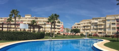 A swimming pool surrounded by a well-maintained lawn, palm trees, and multi-story buildings with balconies.