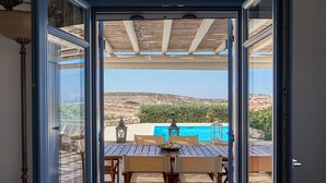 An open door leading to an outdoor patio with a wooden table and chairs, a view of the sea, and a clear sky. - Villa with private wind-sheltered pool, fab seaviews, walking distance to Beach (Paros)