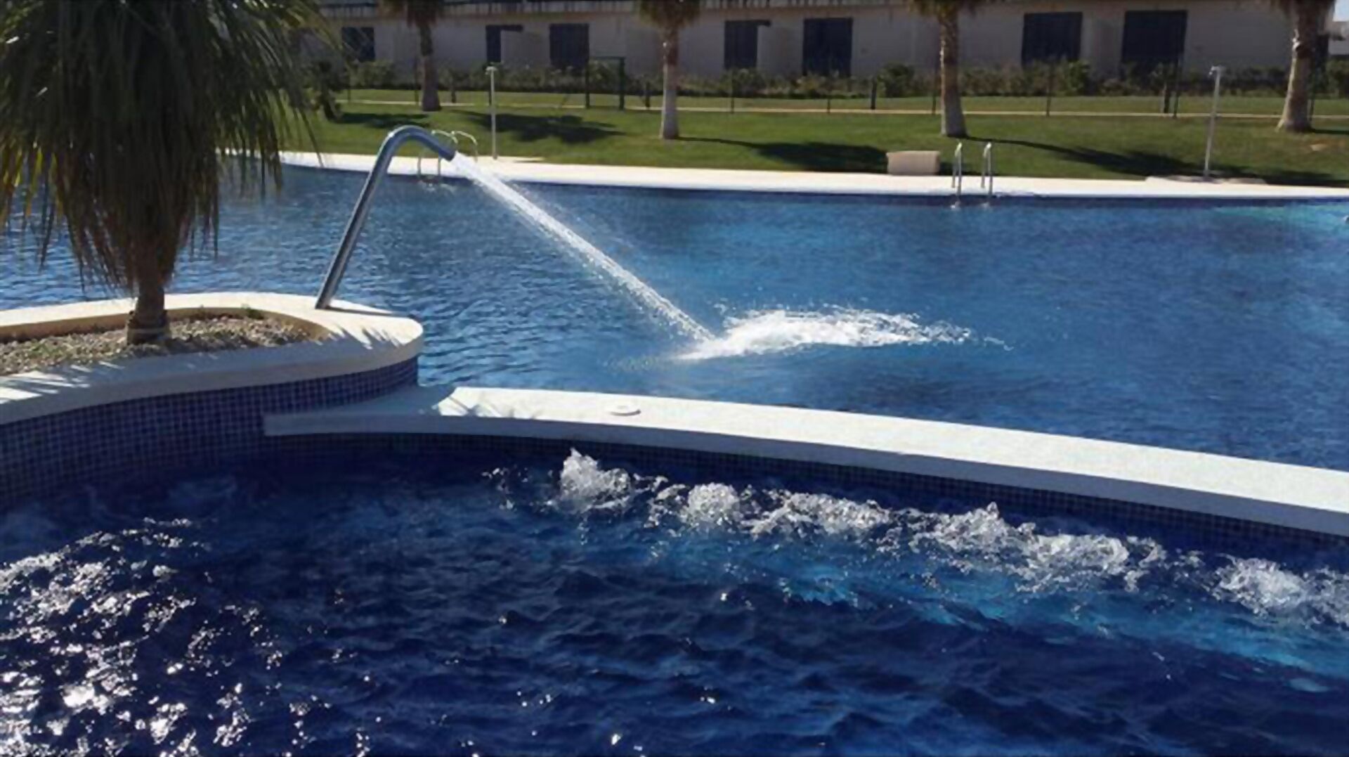 A swimming pool with a water feature, surrounded by a landscaped area with palm trees and a building in the background.
