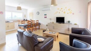 Una sala moderna con un sofá, sillones, una mesa de centro y un televisor. Hay una zona de comedor con sillas y una mesa, y una cocina al fondo.
