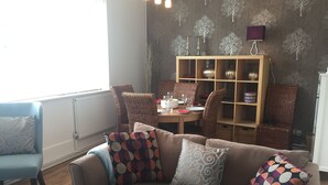 Una sala de estar acogedora con un sofá marrón, cojines estampados, sillas de mimbre, una mesa de comedor con platos y vasos, y una estantería con objetos decorativos.