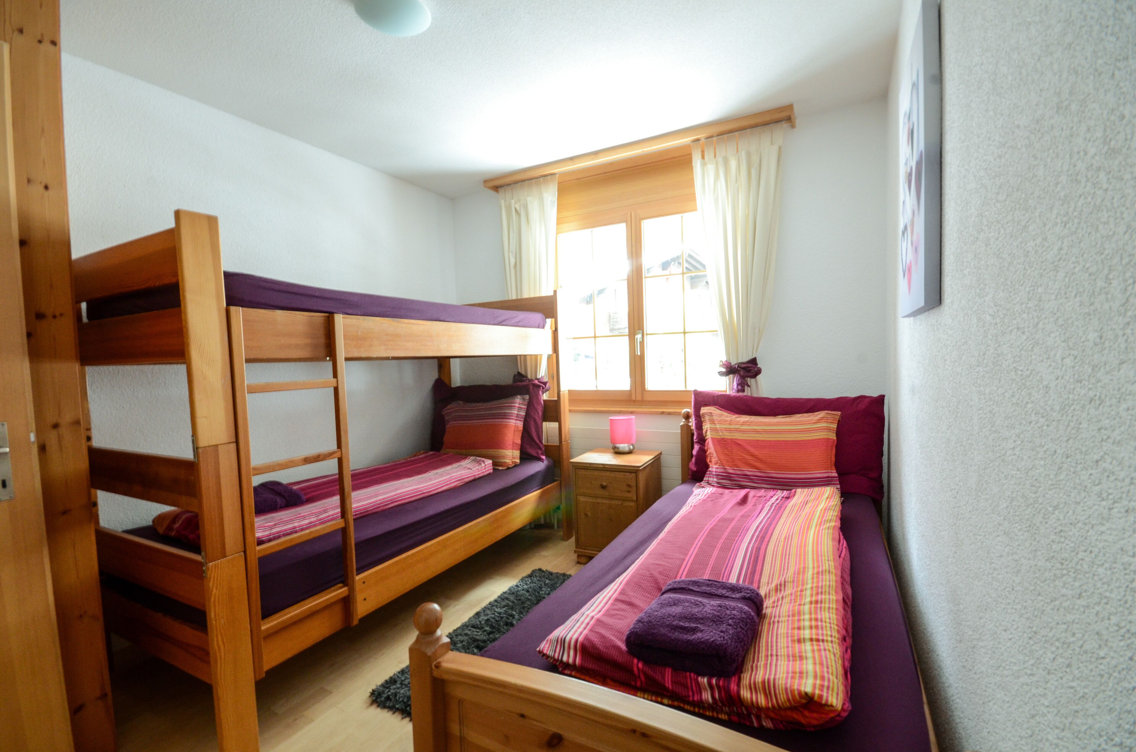 A room with two bunk beds, a small bedside table with a lamp, and a window with curtains.