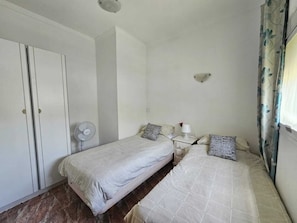 A room with two beds, a wardrobe, a bedside table with a lamp, and a wall-mounted light fixture.