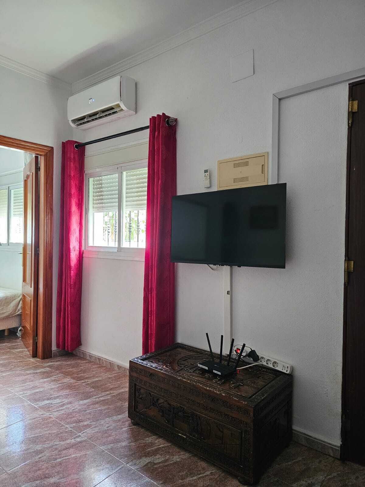 A room with a flat-screen TV mounted on the wall, a wooden chest of drawers, and a window with blinds and red curtains.