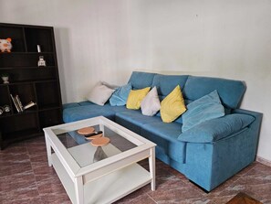 A living room with a blue sofa, a glass coffee table, and a black bookshelf.