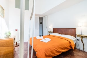 A hotel room with a bed, bedside lamps, a wooden dresser, and a door leading to another room.