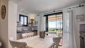 Un coin repas avec une table et des chaises blanches, une cuisine aux carreaux bleus et blancs, et une vue à travers une porte vitrée.