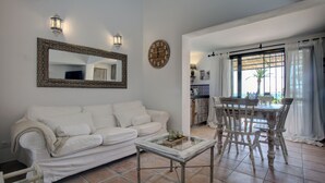Un salon avec un canapé blanc, une table basse en verre, une table à manger en bois avec des chaises, un miroir avec un cadre décoré et une horloge murale.