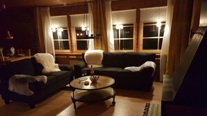 A cozy living room with a dark green sofa, a white furry armchair, and a round coffee table. There are two windows with curtains, a hanging light fixture, and a piano in the corner. - Näset 4 Green Cabin in Småland with boat (Vena)