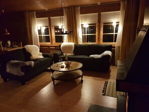 Uma sala de estar aconchegante com um sofá verde escuro, uma poltrona branca e fofa e uma mesa de centro redonda. Há duas janelas com cortinas, uma luminária pendente e um piano no canto.