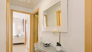 Une chambre d’hôtel moderne avec une porte en bois, un canapé blanc, une petite table sur laquelle se trouvent un bol et une plante, et un grand miroir accroché au mur.